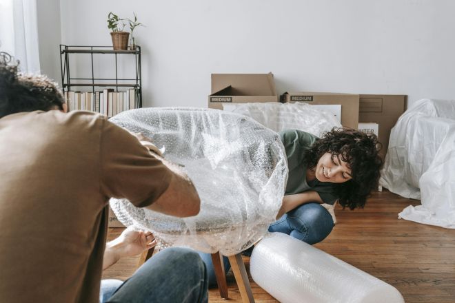 A couple carefully packing chairs with bubble wrap in a sunlit room during a move.