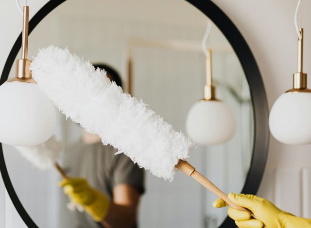 A person cleaning an elegant bathroom mirror with a fluffy duster and yellow gloves.